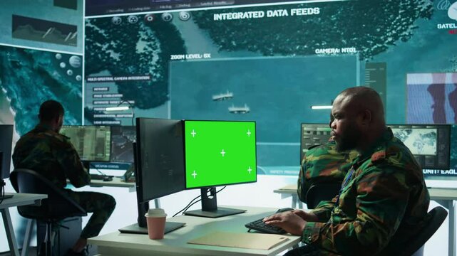 Military specialist monitors satellite data next to green screen display, working at command post with high tech gear to support army defense operations. Coordinates strategic missions. Camera B.
