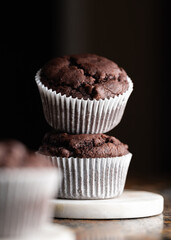 Two stacked chocolate muffins in paper cups on a marble stand on a dark background. Front view of breakfast biscuit cakes.