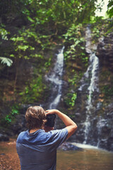 Fototapeta premium Man, camera and photographer by waterfall in nature for photography, rainforest and environment picture of travel. Back, male person and digital gear for creative adventure, exploring and memory
