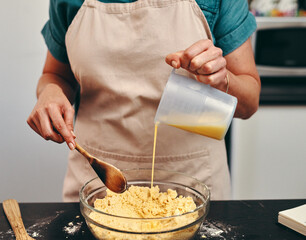 Baking, bowl and hands of chef in kitchen with wooden spoon, pouring wet mix as ingredients for...