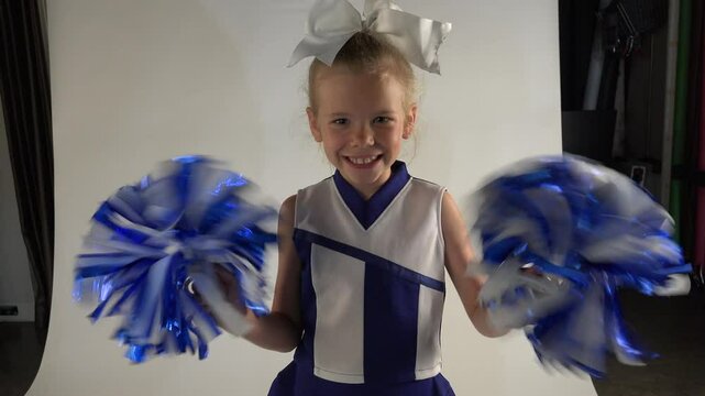 Little girl cheerleader wearing a blue and white cheerleading outfit, shaking her pom poms with glee