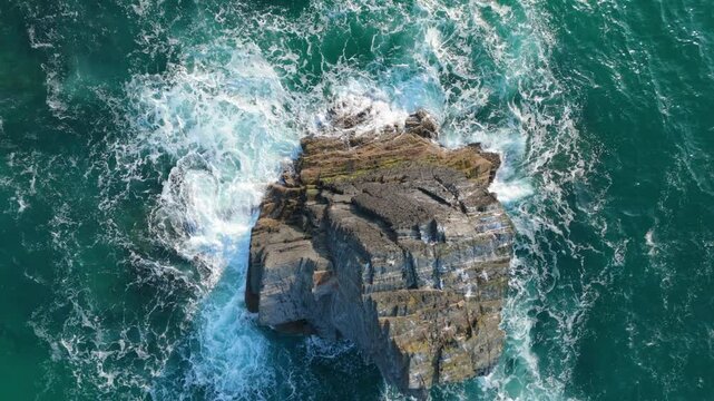 Top down aerial riser over prominent seastack in ocean pummelled with waves