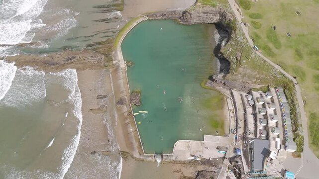 Aerial fly out of Bude Sea Pool in Bude, Cornwall, showcasing the man-made pool integrated into the natural coastline with waves crashing nearby