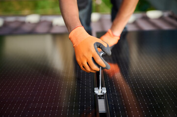 Worker building photovoltaic solar panel system on rooftop of house. Close up of man engineer in gloves installing solar module with help of hex key outdoors. Alternative and renewable energy.