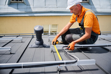 Worker building solar panel system on rooftop of house for generating electricity through photovoltaic effect. Man using ruler to measure mounting equipment for precise installation.