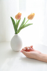 Hands with delicate nail art displayed beside a vase with tulips on a sunlit table.