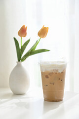 Iced coffee in a plastic cup with whipped cream, placed beside a vase with orange tulips on a sunny day.