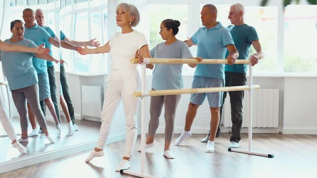 Positive old female attendee of ballet courses performing choreography positions standing by training barre with others