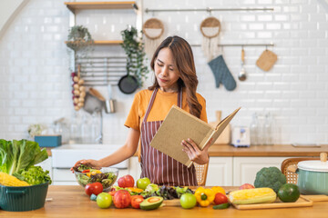 Asian young woman holds a book with online audience or simply enjoying the cooking process, health, fruit, freshness, and organic living, digital recipe platform, eating, healthy at kitchen