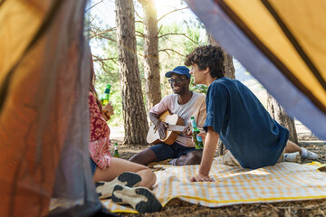 Diverse friends enjoying music and drinks at campsite, multicultural group relaxing in forest,...