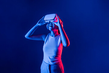 Smart female stand with surrounded by cyberpunk neon light wear VR headset connecting metaverse, futuristic cyberspace community technology. Elegant woman look generated virtual area. Hallucination.