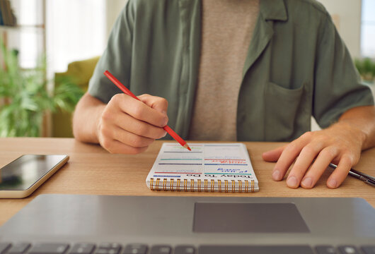 Male college student writing to do list in his planner. Crop shot of young man sitting at table with paper notebook and laptop PC, holding pencil in hand and reading information about upcoming events