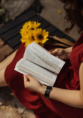 A woman in a burgundy dress sits on a wooden bench and turns the pages of a book in her hands, a...