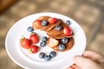 A man's hand holds a plate of sugar-free, gluten-free green buckwheat fritters with strawberries and blueberries . Healthy and delicious pancakes.