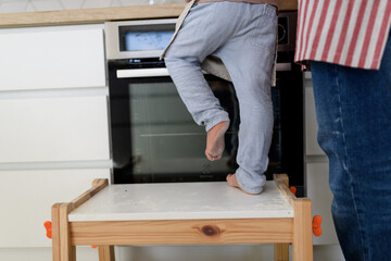Fototapeta premium Boy learning how to make pancakes, standing on chair by stove. Parents and children in kitchen, preparing pancake batter, spending weekend day indoors.
