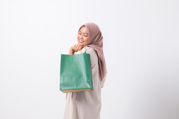 Portrait of cheerful Asian hijab woman in casual suit holding and showing colorful paper shopping bags. Shopaholic girl and discount buying concept. Isolated image on white background