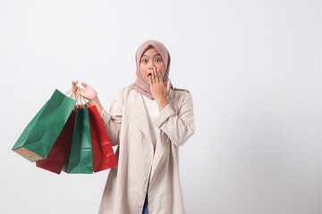 Portrait of shocked Asian hijab woman in casual suit holding paper shopping bags while covering mouth. Shopaholic girl and discount buying concept. Isolated image on white background