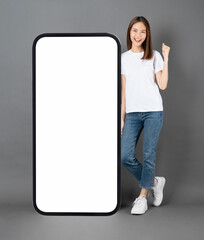 Cheerful and beautiful Asian woman stands beside a large smartphone mockup with a blank screen against a grey background.