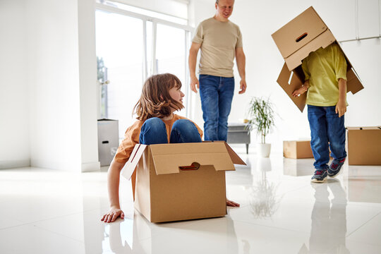 Siblings playing with cardboard boxes near father in empty room of new apartment