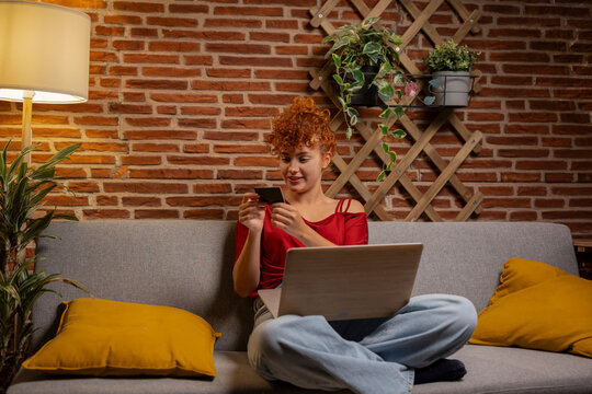 Curly hair young woman doing online shopping using credit card and sitting on sofa with laptop at home