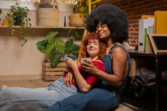 Young woman with eyes closed leaning on friend sitting on floor having coffee at home
