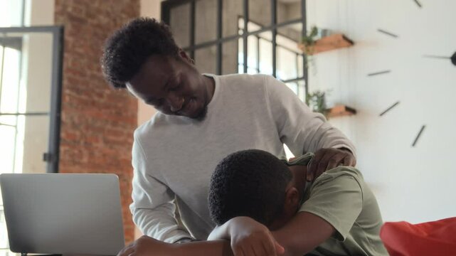 African American boy returns from school sad and upset, sits down at table near working father, put head on desk. Black skinned man type on laptop, son tired after classes, dad supports and calms him