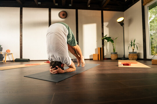 Young man practicing a headstand yoga pose indoors in a serene, well-lit room with wooden beams and large windows.