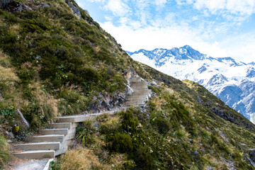 A winding staircase path ascends a green mountain slope with snow-capped peaks in the background, under a partly cloudy sky, offering a serene hiking trail.