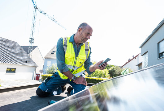 Technician with smart phone installing solar panel on house rooftop