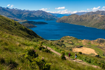 Two hikers stand on a trail overlooking a beautiful lake surrounded by mountains. The clear blue sky and lush greenery enhance the breathtaking view, creating a sense of adventure and exploration.