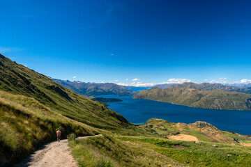 A lone hiker walks along a mountain path overlooking a vast, tranquil lake surrounded by rolling...