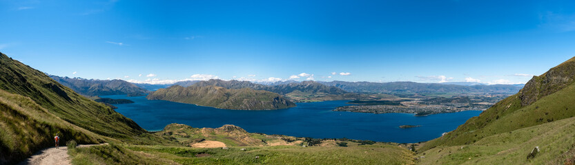Fototapeta premium A panoramic view of a lake surrounded by mountains, with a hiker on a trail in the foreground, under a clear blue sky.