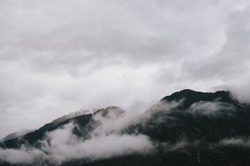 A serene landscape of a mountain range enveloped in mist, with dark green forests at the base, under a cloudy, overcast sky.