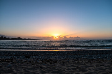 The sun sets over a rocky beach, casting a warm glow on the water and stones. The scene captures the tranquility and natural beauty of the coastal landscape at dusk.
