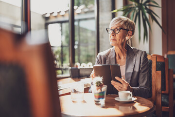 Mature woman is sitting in cafe and relaxing. She is drinking coffee and using digital tablet.