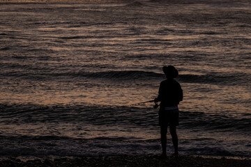 Fototapeta premium A silhouette of a man fishing by the ocean at sunset, capturing a serene mood. The dark figure contrasts against the rippling water illuminated by fading light, emphasizing tranquility.