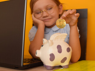 A unique child, a little cute girl sitting near a laptop puts Bitcoin coins into a piggy bank. A child learns from childhood to earn cryptocurrency. The child is a young genius, businessman.
