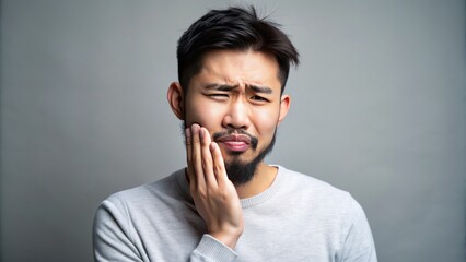 Concerned young Asian man with a beard stands on a gray background clutching his painful lower jaw with both hands.
