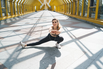 young Caucasian woman performs deep side lunge stretch in urban setting. Dressed in sports bra and leggings, smiles focuses on flexibility during morning workout, enjoying vibrant and active lifestyle