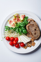 Breakfast plate with bread, tomatoes, cheese, arugula, nuts and figs on white background.