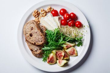 Breakfast plate with bread, tomatoes, cheese, arugula, nuts and figs on white background.