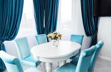 Bright Dining Room with White Table and Blue Chairs.Spacious dining area with a white table, blue velvet chairs, and a floral centerpiece, illuminated by natural light. 