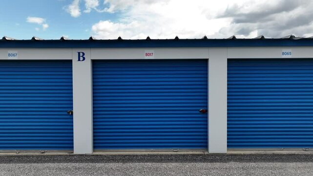 Row of blue storage unit doors in a self-storage facility. Each unit labeled with numbers and letters. Simple, secure storage solution. Aerial.