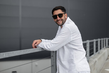 Happy handsome guy with fashion sunglasses in white clothes with shirt stands near a modern building in the city