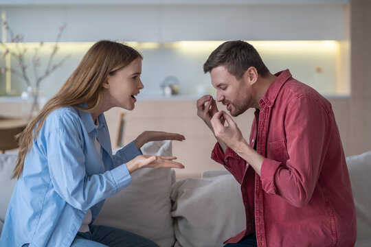 Shouting romantic couple sits face to face on couch, actively gesticulating and arguing using sign language at home, expressing emotions and perspectives through their animated hand movements.
