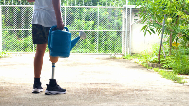 An elderly man with a prosthetic leg carries a bucket of water to water his vegetables in his garden in front of his house. The background is blurred.
