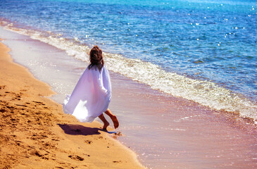 Happy little girl in a pink pareo running along the beach back to the camera in summer