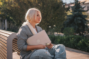A woman is sitting on a bench with a laptop in front of her. She is smiling and she is enjoying her...