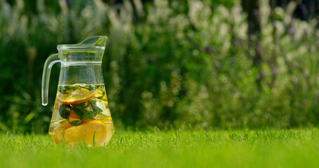 Glass pitcher with lemon and mint lemonade placed on a green lawn in a sunny garden.