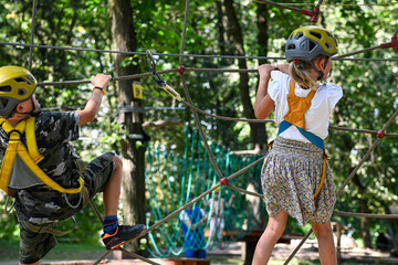 Two children wearing safety harnesses and helmets navigating a rope course in a park